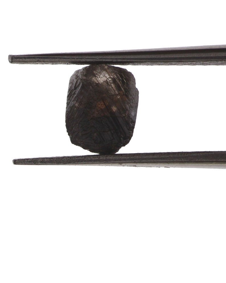 Puede incluir: Una piedra preciosa facetada de color gris oscuro sostenida por unas pinzas. La piedra preciosa es rugosa y tiene un aspecto natural sin pulir.