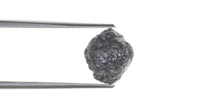 Pode incluir: Um diamante bruto cinza escuro, preso por uma pin&ccedil;a contra um fundo branco. A gema apresenta uma superf&iacute;cie irregular e texturizada, destacando sua forma natural e apar&ecirc;ncia n&atilde;o lapidada.