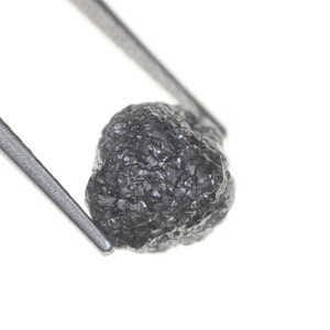 Pode incluir: Um diamante negro bruto e n&atilde;o lapidado, segurado por pin&ccedil;as prateadas contra um fundo branco. O diamante tem uma superf&iacute;cie texturizada e uma forma irregular, mostrando sua forma natural. Esta imagem destaca a beleza bruta da gema.