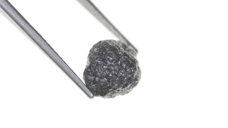 Pode incluir: Um diamante negro bruto e n&atilde;o lapidado, segurado por pin&ccedil;as prateadas contra um fundo branco. O diamante tem uma superf&iacute;cie texturizada e uma forma irregular, mostrando sua forma natural. Esta imagem destaca a beleza bruta da gema.