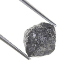 Pode incluir: Um diamante bruto cinza escuro, segurado por pin&ccedil;as prateadas contra um fundo branco. O diamante tem uma superf&iacute;cie texturizada e uma forma irregular, mostrando sua forma natural. As pin&ccedil;as est&atilde;o posicionadas para exibir o diamante.