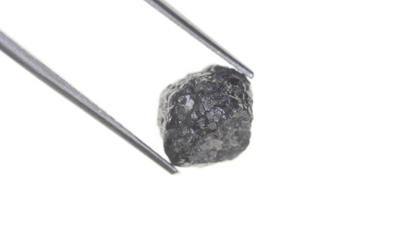 Pode incluir: Um diamante bruto cinza escuro, segurado por pin&ccedil;as prateadas contra um fundo branco. O diamante tem uma superf&iacute;cie texturizada e uma forma irregular, mostrando sua forma natural. As pin&ccedil;as est&atilde;o posicionadas para exibir o diamante.