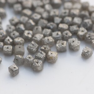 May include: A close-up of a pile of small, gray, square-shaped diamonds. The diamonds have a rough, textured surface and a small hole drilled through the center.