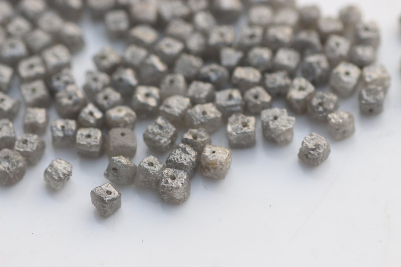 May include: A close-up of a pile of small, gray, square-shaped diamonds. The diamonds have a rough, textured surface and a small hole drilled through the center.