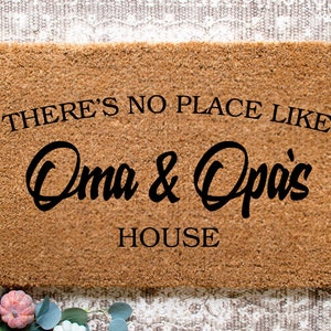 May include: A rectangular doormat with the text "THERE'S NO PLACE LIKE Oma & Opa's HOUSE" in black script. The mat is a natural brown color and is likely made of coir. Decorative elements are visible in the corners.