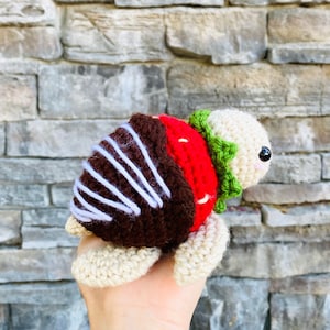 May include: A crocheted strawberry turtle toy with a brown shell, white stripes, and green leaves. The turtle is holding its head up and looking to the right.