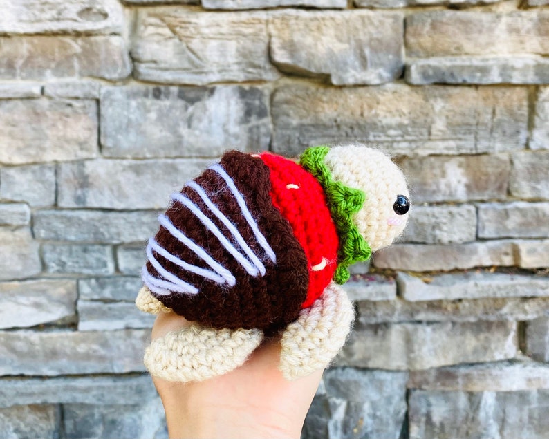 Peut inclure: Une tortue en crochet en forme de fraise avec une carapace brune, des rayures blanches et des feuilles vertes. La tortue l&egrave;ve la t&ecirc;te et regarde vers la droite.