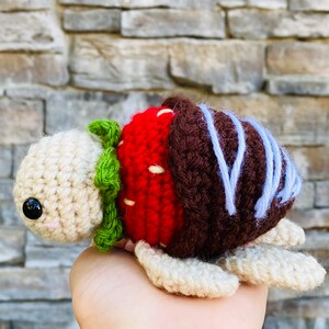 Peut inclure: Un jouet en peluche en crochet en forme de tortue avec une carapace de fraise rouge et blanche, une carapace de chocolat marron et des d&eacute;tails de gla&ccedil;age bleu.