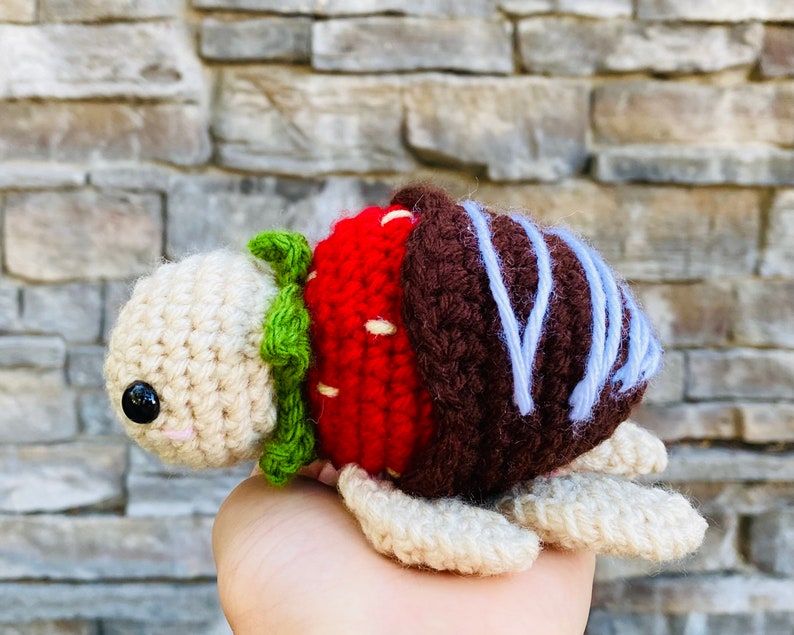 Peut inclure: Un jouet en peluche en crochet en forme de tortue avec une carapace de fraise rouge et blanche, une carapace de chocolat marron et des d&eacute;tails de gla&ccedil;age bleu.