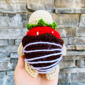 Peut inclure: Un jouet en peluche en crochet en forme de tortue avec une fraise rouge et verte sur le dos. La fraise est faite de laine et a une garniture en chocolat brun.