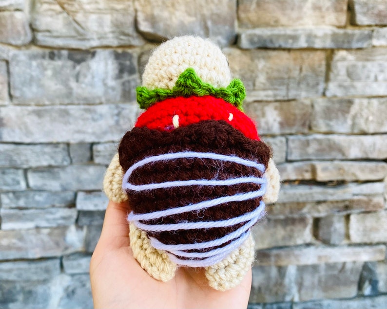 Peut inclure: Un jouet en peluche en crochet en forme de tortue avec une fraise rouge et verte sur le dos. La fraise est faite de laine et a une garniture en chocolat brun.