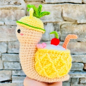 Pode incluir: Um caracol de crochê usando um chapéu de abacaxi e carregando um abacaxi amarelo nas costas. O caracol tem um coração rosa em sua concha e uma cereja vermelha no topo do abacaxi.
