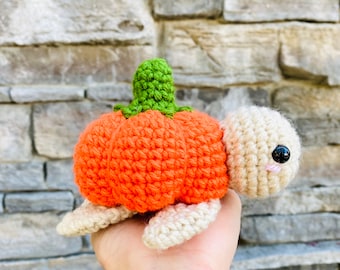 May include: A crocheted turtle with an orange pumpkin shell. The turtle is made of beige yarn and has black eyes. The pumpkin shell has a green stem.