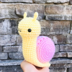 May include: A crocheted snail toy with a yellow body and a pink shell. The snail has black eyes and a small, stitched smile.