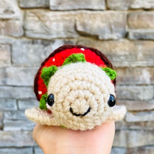 Peut inclure: Un jouet de tortue en crochet en forme de fraise avec une carapace rouge et verte et des yeux noirs. La tortue sourit et a un corps beige.