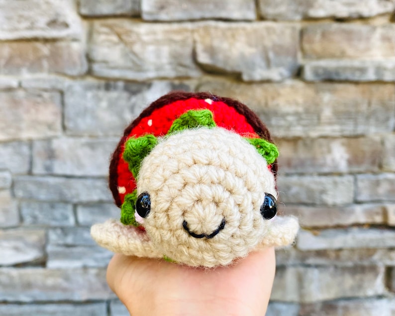 Peut inclure: Un jouet de tortue en crochet en forme de fraise avec une carapace rouge et verte et des yeux noirs. La tortue sourit et a un corps beige.