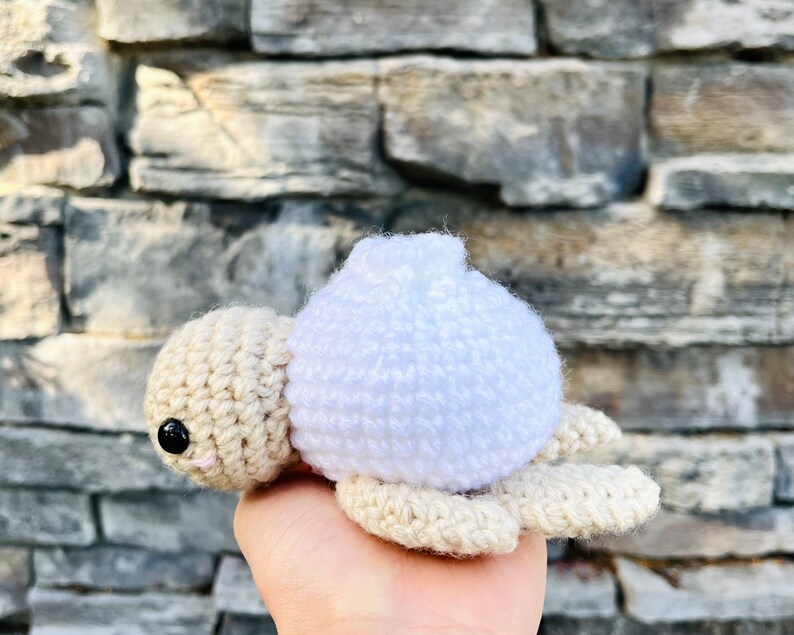 Op de afbeelding: Een gehaakt zeeschildpad speelgoed met een witte schelp en een beige lichaam. De schildpad zit op een hand.