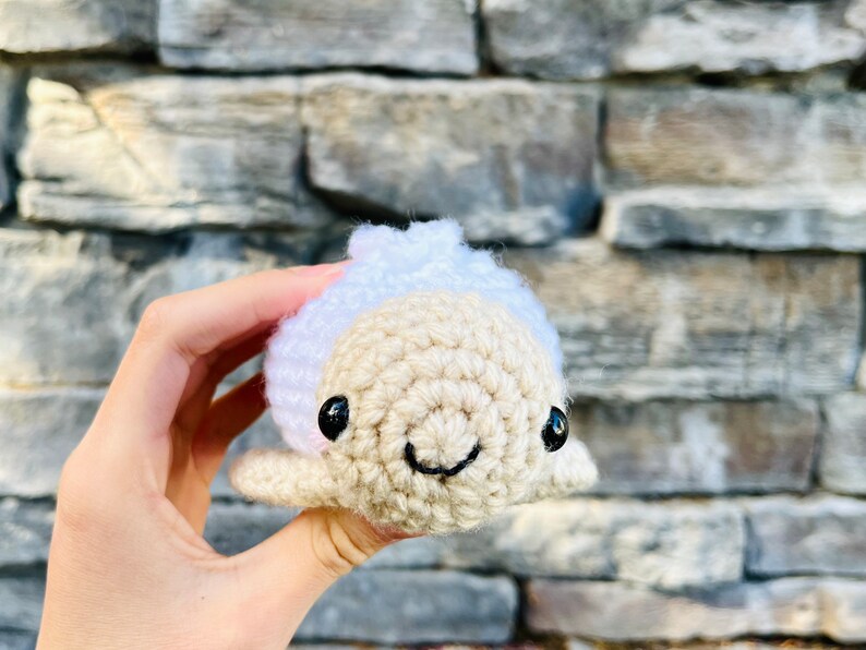 Op de afbeelding: Een gehaakt pluche speelgoed in de vorm van een dolfijn met een wit hoofd en een beige lichaam. De dolfijn heeft zwarte ogen en een glimlachende mond.