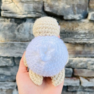 Op de afbeelding: Een gehaakt speelgoed in de vorm van een schildpad met een witte schild en een beige lichaam. De schildpad is met zijn rug naar de camera gekeerd.