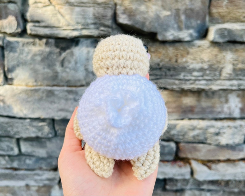 Op de afbeelding: Een gehaakt speelgoed in de vorm van een schildpad met een witte schild en een beige lichaam. De schildpad is met zijn rug naar de camera gekeerd.