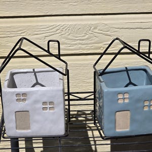 May include: Two ceramic house-shaped planters, one white and one blue, each with a door and window cutouts. They are suspended in black metal frames, resembling miniature houses. The planters are designed for small plants or decorative use.