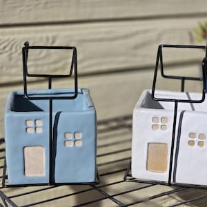May include: Two ceramic house-shaped planters, one light blue and one white, each with a black metal frame. The planters have square windows and a rectangular door. The planters are sitting on a black metal shelf.