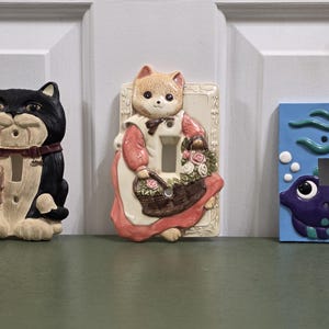 May include: Three decorative light switch covers. One features a black and white cat with a mouse, another a cat with a basket of flowers, and the last a blue cover with a purple fish and bubbles.
