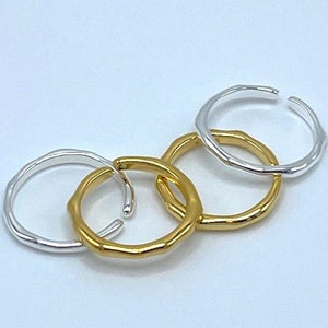 May include: Four simple, minimalist rings, two gold and two silver, are stacked on a white surface. The rings are all smooth and have a slightly irregular shape.