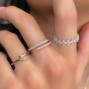 May include: Three delicate rings with clear stones on a finger. The rings are made of gold and have a simple design. The ring on the middle finger is a thin band with a row of small stones. The ring on the index finger is a thin band with a row of small stones. The ring on the pinky finger is a thin band with a row of small stones.