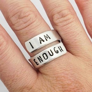 May include: A silver ring with the words "I AM ENOUGH" engraved on it. The ring is worn on a finger with light blue nail polish.