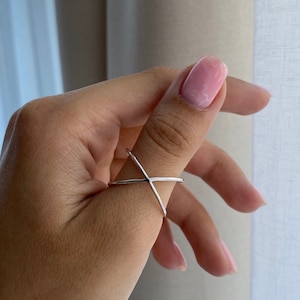 May include: A silver ring with a simple, geometric design. The ring is made of two thin bands that cross each other in the center, creating an X shape.