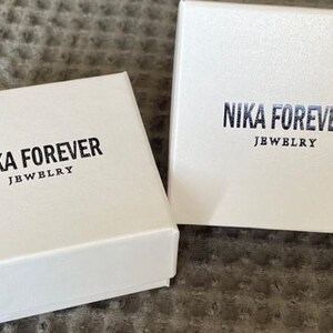 May include: Two white boxes with the text "NIKA FOREVER JEWELRY" printed on them. The boxes are on a brown textured surface.
