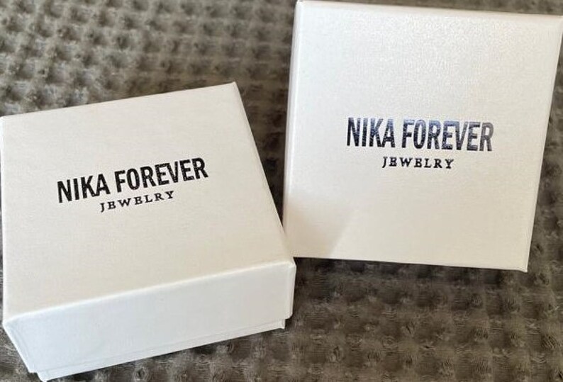 May include: Two white boxes with the text "NIKA FOREVER JEWELRY" printed on them. The boxes are on a brown textured surface.