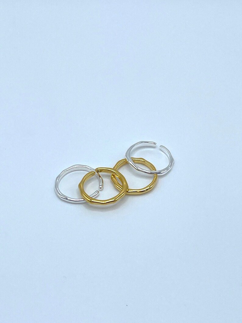 May include: Four simple, minimalist rings, two gold and two silver, are stacked on a white surface. The rings are all smooth and have a slightly irregular shape.