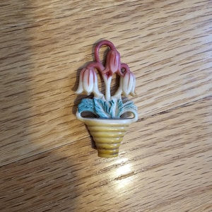 May include: A small, decorative pin featuring a yellow flower pot with a ribbed design. The pot holds three stylized flowers with white petals and red-orange tips, and green leaves. The pin is made of a light-colored material.