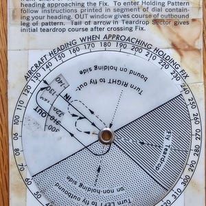 May include: A vintage holding pattern computer with a circular dial and text. The dial features markings for aircraft heading and holding fix, with instructions for inbound and outbound courses. The device is designed for aviation navigation.
