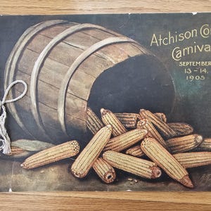 May include: Vintage poster art for the Atchison Corn Carnival, featuring a wooden barrel spilling out ears of corn. The poster is tied with a string and displays the event's name and date: September 13-14, 1905.