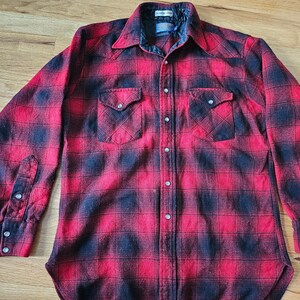 May include: A long-sleeve button-down shirt featuring a red and black buffalo plaid pattern. The shirt includes two chest pockets with pointed flaps, a pointed collar, and silver-toned snap buttons down the front placket and on the cuffs.