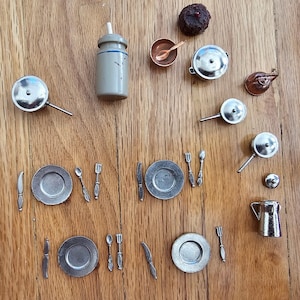 May include: A collection of miniature dollhouse kitchenware, including pots, pans, plates, cutlery, and a small pitcher. The items are made of metal and are arranged on a wooden surface. A small, dark-colored food item is also present.