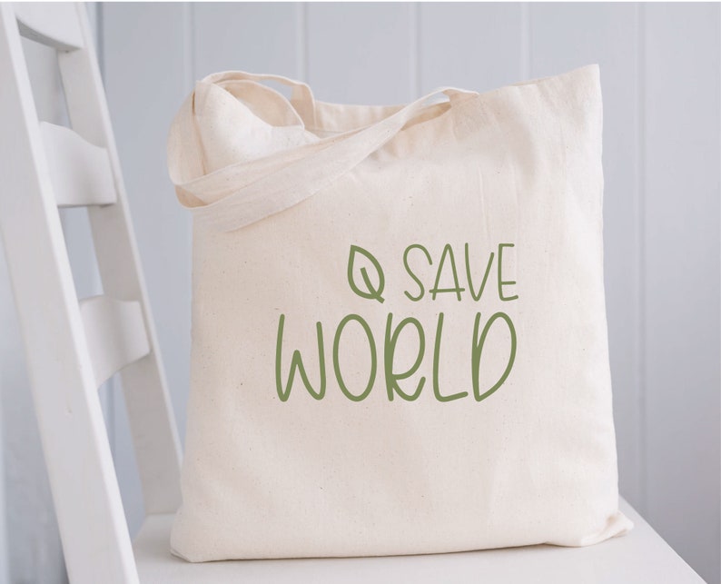 Pode incluir: Uma bolsa tote de lona branca com texto verde que diz "Q SAVE WORLD" e um pequeno desenho de folha verde.