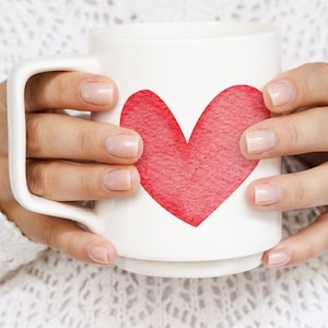 K&ouml;nnte beinhalten: Eine wei&szlig;e Keramiktasse mit einem roten Herzdesign. Die Tasse wird von den H&auml;nden einer Person gehalten.