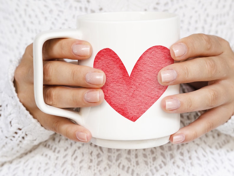 K&ouml;nnte beinhalten: Eine wei&szlig;e Keramiktasse mit einem roten Herzdesign. Die Tasse wird von den H&auml;nden einer Person gehalten.