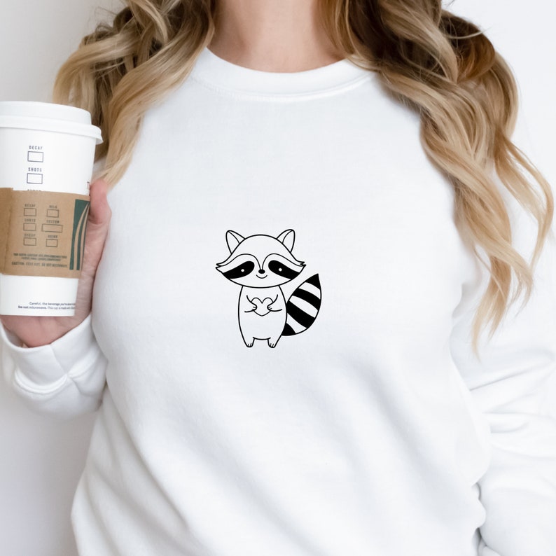 May include: White sweatshirt with a black and white cartoon raccoon holding a heart. The raccoon has a black nose and eyes.