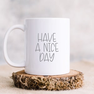 Pode incluir: Caneca de cer&acirc;mica branca com uma mensagem escrita &agrave; m&atilde;o em preto que diz "Have a nice day". A caneca est&aacute; sentada em uma fatia de madeira com musgo.