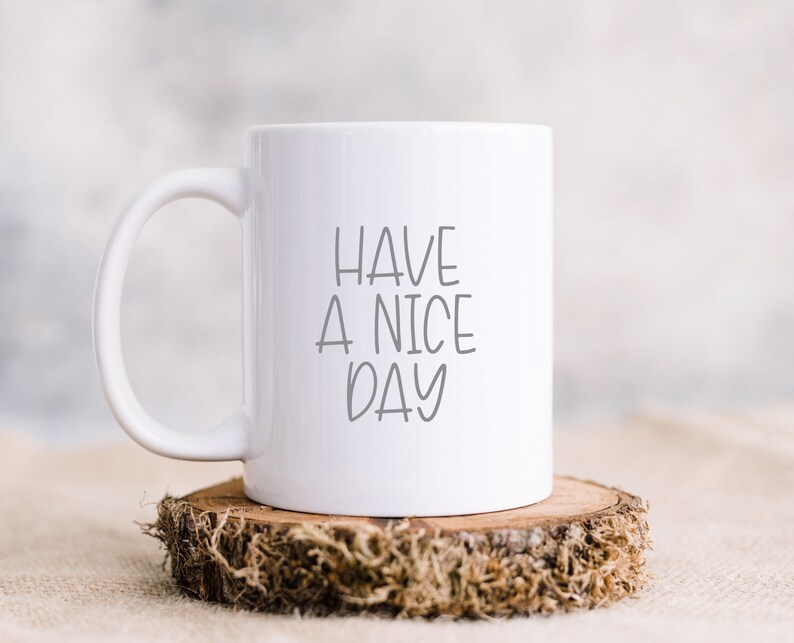 Pode incluir: Caneca de cer&acirc;mica branca com uma mensagem escrita &agrave; m&atilde;o em preto que diz "Have a nice day". A caneca est&aacute; sentada em uma fatia de madeira com musgo.