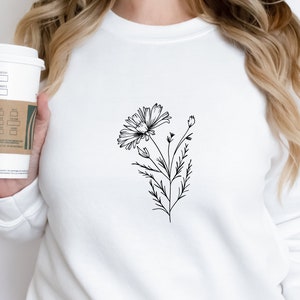 Puede incluir: Una sudadera blanca con un dibujo de l&iacute;nea negra de una flor en la parte delantera.