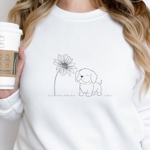 May include: White sweatshirt featuring a black and white line drawing of a smiling puppy next to a sunflower. The design is simple and whimsical, perfect for dog lovers. The sweatshirt is a comfortable, casual style.