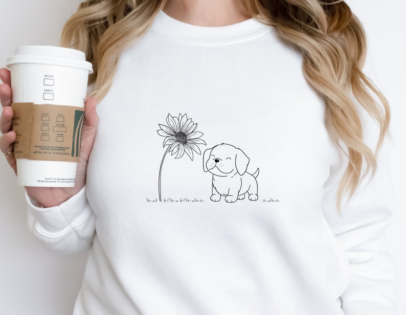 May include: White sweatshirt featuring a black and white line drawing of a smiling puppy next to a sunflower. The design is simple and whimsical, perfect for dog lovers. The sweatshirt is a comfortable, casual style.