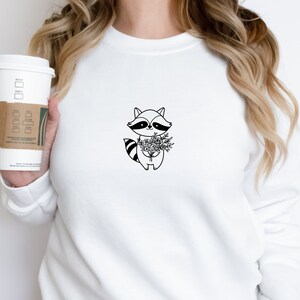 May include: White sweatshirt with a black line drawing of a raccoon holding a bouquet of flowers.