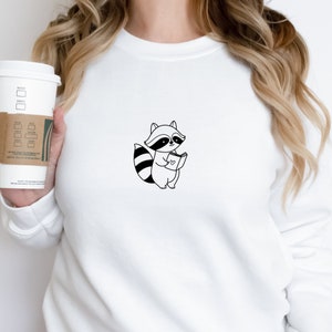 May include: White sweatshirt with a black and white illustration of a raccoon reading a book with a heart on the cover.